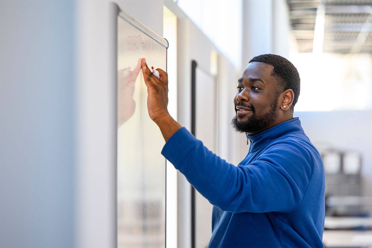 A person is standing in a bright room, in front of a dry erase marker board. There is writing on the board and the person is using their hand to point to or erase a segment of the writing.